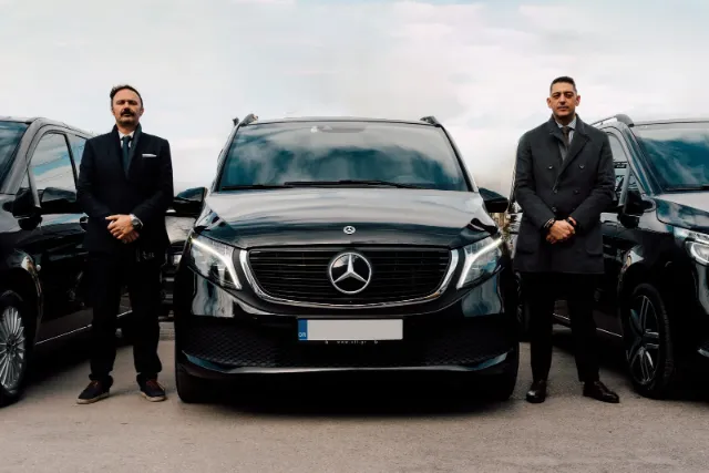 Two chauffeurs in dark suits beside a black Mercedes-Benz V-Class van